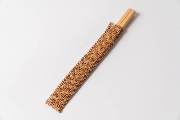 約1万年前の縄文時代からの手技の流れを汲む伝統的な織物「しな織」は、しなの木の樹皮を繊維にした、世界でも稀な自然布。軽くて水に強く、しなやか。

昔は網や袋に使われていたこの素材を、現代の暮らしに合う箸入れとして仕立てました。

通気性が高く、食事のたびに手に取る道具として最適。素朴な風合い、自然な柄の浮き出し、時間とともに深まる色味と柔らかさ。日常に溶け込む一方で、しな織の美しい存在感も楽しめます。

白い木綿糸で施したステッチは、100年以上前の「しな織の蒸し布」に由来。古布の縫い目をオマージュしたデザインが、文化と暮らしの融合を感じさせてくれます。